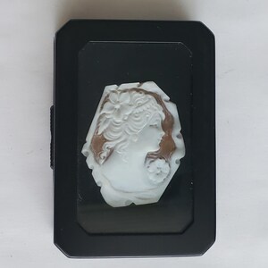 May include: A black rectangular jewelry box with a white cameo of a woman's profile. The cameo is set against a brown background and features intricate details of the woman's hair and floral accents.
