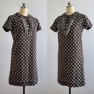 May include: A brown and white polka dot dress with a white ruffled collar and a white beaded necklace. The dress has short sleeves and a straight silhouette.