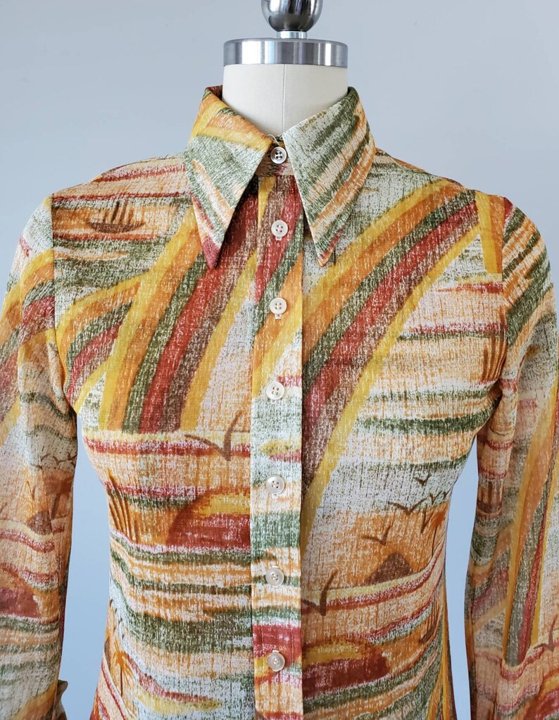 May include: A long-sleeved button-down shirt with a colorful abstract print in shades of orange, yellow, green, and brown. The shirt has a pointed collar and a button-up front.