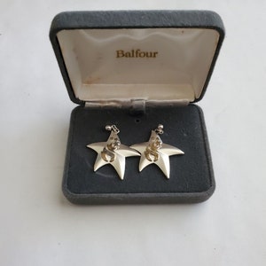 Könnte beinhalten: Ein Paar silberne sternförmige Ohrringe mit einem goldfarbenen Design in jedem Stern. Die Ohrringe befinden sich in einer grauen Samtbox mit dem Wort "Balfour" auf der Innenseite des Deckels.