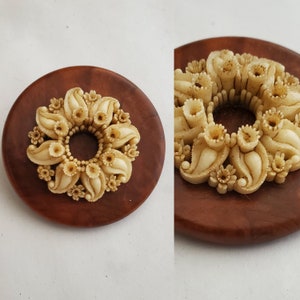 May include: A white, carved floral design brooch on a brown wooden base. The brooch is in the shape of a circle with a central hole and features intricate details of leaves and flowers.