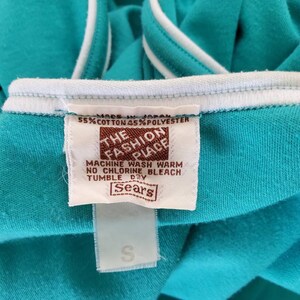 May include: A turquoise blue t-shirt with white trim. The tag reads "The Fashion Place" and includes care instructions. The tag also says "Made in Japan" and "55% Cotton 45% Polyester". The tag is brown with white text. The t-shirt is labeled "Sears".