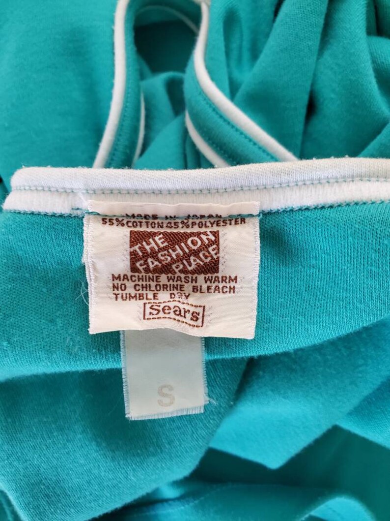 May include: A turquoise blue t-shirt with white trim. The tag reads "The Fashion Place" and includes care instructions. The tag also says "Made in Japan" and "55% Cotton 45% Polyester". The tag is brown with white text. The t-shirt is labeled "Sears".