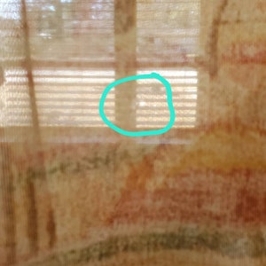 May include: A close-up of a sheer curtain with a window behind it. The window has blinds and the curtain has a pattern of brown, orange, and green.