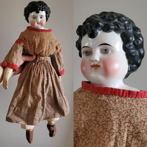 May include: Antique porcelain doll with black curly hair, blue eyes, and rosy cheeks. The doll is wearing a brown and white floral dress with red trim and brown leather shoes.