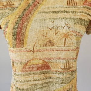May include: A beige, brown, and green patterned shirt with a short sleeve and a crew neck. The pattern features a stylized landscape with palm trees, birds, and hills.