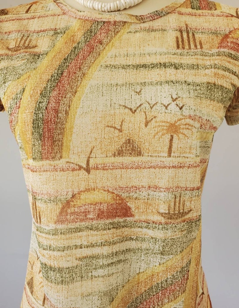 May include: A beige, brown, and green patterned shirt with a short sleeve and a crew neck. The pattern features a stylized landscape with palm trees, birds, and hills.
