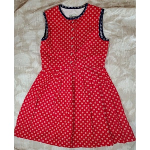 May include: A sleeveless red dress with a white floral pattern. The dress features a button-down front, a fitted waist, and a flared skirt. The neckline and armholes are trimmed with navy blue fabric.