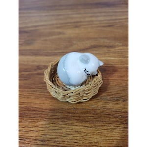 May include: A small, ceramic cat figurine, curled up in a woven basket. The cat is white with gray markings and has closed eyes, suggesting sleep. The basket is made of light brown woven material.