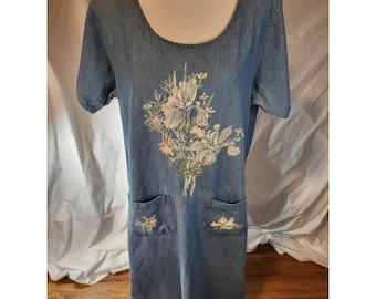 May include: A light blue denim dress with short sleeves and a scoop neckline. The dress features a floral embroidery design on the front and two front pockets. The floral design includes white, light blue, and green colors.