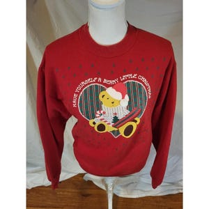 Puede incluir: Sudadera roja de Navidad con un oso de peluche con gorro de Papá Noel dentro de un diseño en forma de corazón. El texto "Have yourself a berry little Christmas" está arqueado alrededor del corazón. Pequeños árboles de Navidad verdes están dispersos.
