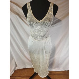 May include: A vintage ivory-colored slip dress on a black mannequin. The bodice features a delicate floral lace design with a V-neckline. The skirt is a smooth, flowing fabric, and the hemline is trimmed with lace.