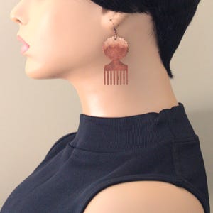 May include: Copper-colored earrings in the shape of a comb and a circular design. The earrings are hanging from a mannequin wearing a black top and a black wig with bangs. The earrings are a unique accessory.
