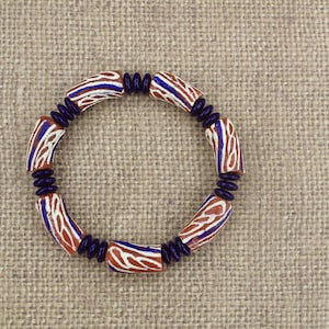 Puede incluir: Un brazalete hecho de cuentas cilíndricas marrones, blancas y azules. Las cuentas tienen un patrón de rayas.