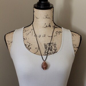 May include: A copper wire wrapped pendant necklace on a black cord. The pendant is a spiral shape and is hanging from a mannequin wearing a white tank top and a colorful patterned skirt.