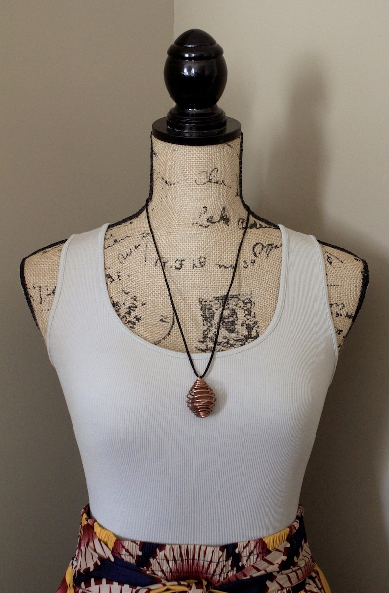 May include: A copper wire wrapped pendant necklace on a black cord. The pendant is a spiral shape and is hanging from a mannequin wearing a white tank top and a colorful patterned skirt.