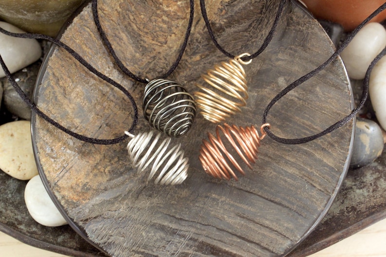 May include: Four wire-wrapped pendants in different colors, including silver, gold, and copper, are displayed on a wooden surface. Each pendant is a spiral shape and hangs from a black cord.