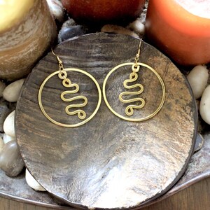 Puede incluir: Un par de pendientes de aro dorados con un diseño ondulado único en el interior de los aros. Los pendientes se exhiben sobre una superficie oscura y texturizada, rodeados de pequeñas piedras y velas, creando una estética cálida. Un accesorio elegante.