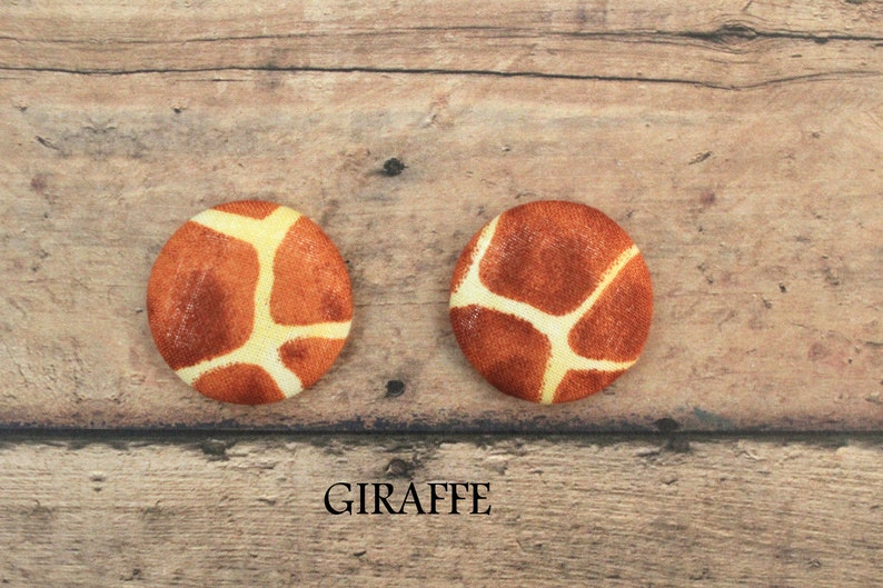 Pu&ograve; includere: Due bottoni rotondi con stampa di giraffa. I bottoni sono marroni con macchie gialle e sono su uno sfondo di legno. Il testo "GIRAFFE" &egrave; stampato sotto i bottoni.