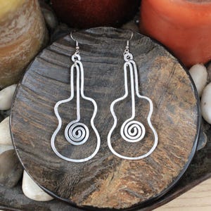 May include: A pair of silver-colored guitar-shaped earrings. Each earring features a wire outline of a guitar with a spiral design at the sound hole. The earrings are displayed on a dark, textured surface, with candles and stones in the background.