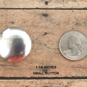 Pu&ograve; includere: Un bottone in metallo argentato di 3 cm di diametro, accanto a una moneta da 25 centesimi di dollaro americano per il confronto delle dimensioni. Bottone piccolo.