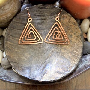 Puede incluir: Un par de pendientes de cobre en forma de triángulo con un diseño en espiral. Los pendientes están sobre una superficie de madera con pequeñas piedras.