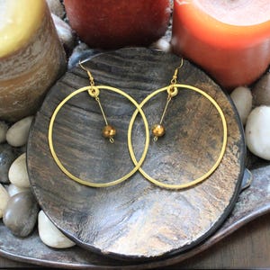May include: Gold hoop earrings with a dangling gold bead. The earrings are displayed on a dark, textured plate, surrounded by white and gray stones, and candles. The earrings have a simple, elegant design.