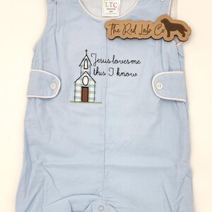 May include: A light blue sleeveless romper featuring a white and blue checkered church embroidery with the text Jesus loves me this I know. The garment has white trim, side button tabs, and a bottom snap closure. A wooden tag reads The Red Lab Co.