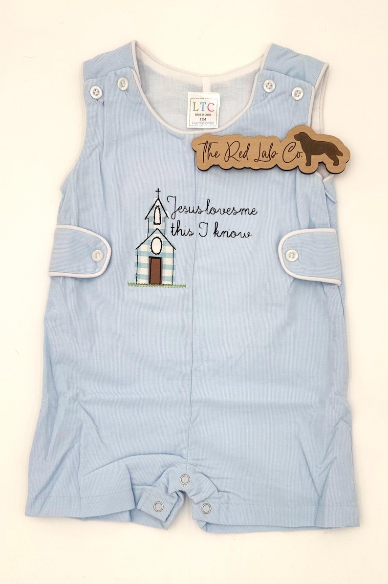 May include: A light blue sleeveless romper featuring a white and blue checkered church embroidery with the text Jesus loves me this I know. The garment has white trim, side button tabs, and a bottom snap closure. A wooden tag reads The Red Lab Co.
