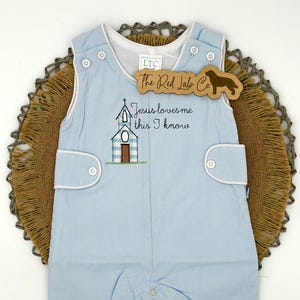 May include: A light blue sleeveless romper with white trim, featuring an embroidered church graphic and the text "Jesus loves me this I know". The garment has button closures at the shoulders and sides, and a snap closure at the bottom hem.
