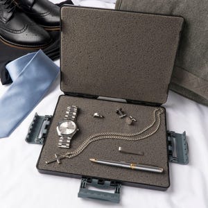Puede incluir: Una maleta de viaje negra con un interior de espuma gris. La maleta está abierta y contiene un reloj plateado, un collar de cadena plateada con un colgante de cruz, un broche de corbata plateado y un bolígrafo plateado.