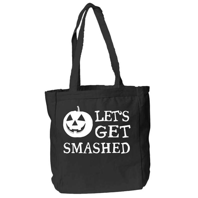 May include: Black tote bag with white text and a jack-o'-lantern graphic. The text reads "LET'S GET SMASHED". The bag has two black handles and is made of a sturdy material, perfect for carrying groceries or other items.