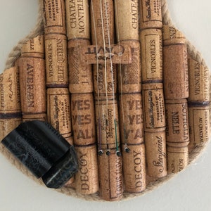 Pu&ograve; includere: Un violino realizzato con tappi di sughero per vino, con corde e un bordo in corda marrone. I tappi hanno varie etichette e testo, tra cui "Chateau Monteles", "Vignobles", "Gillet-Queyrens", "Yes", "Decoy", "Storypoint" e "Bottiglia Nielle".