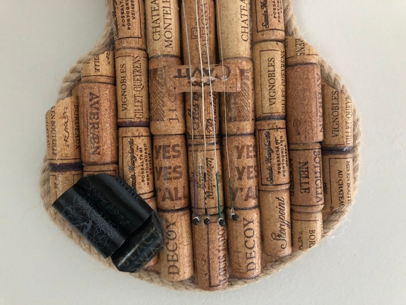 Pu&ograve; includere: Un violino realizzato con tappi di sughero per vino, con corde e un bordo in corda marrone. I tappi hanno varie etichette e testo, tra cui "Chateau Monteles", "Vignobles", "Gillet-Queyrens", "Yes", "Decoy", "Storypoint" e "Bottiglia Nielle".