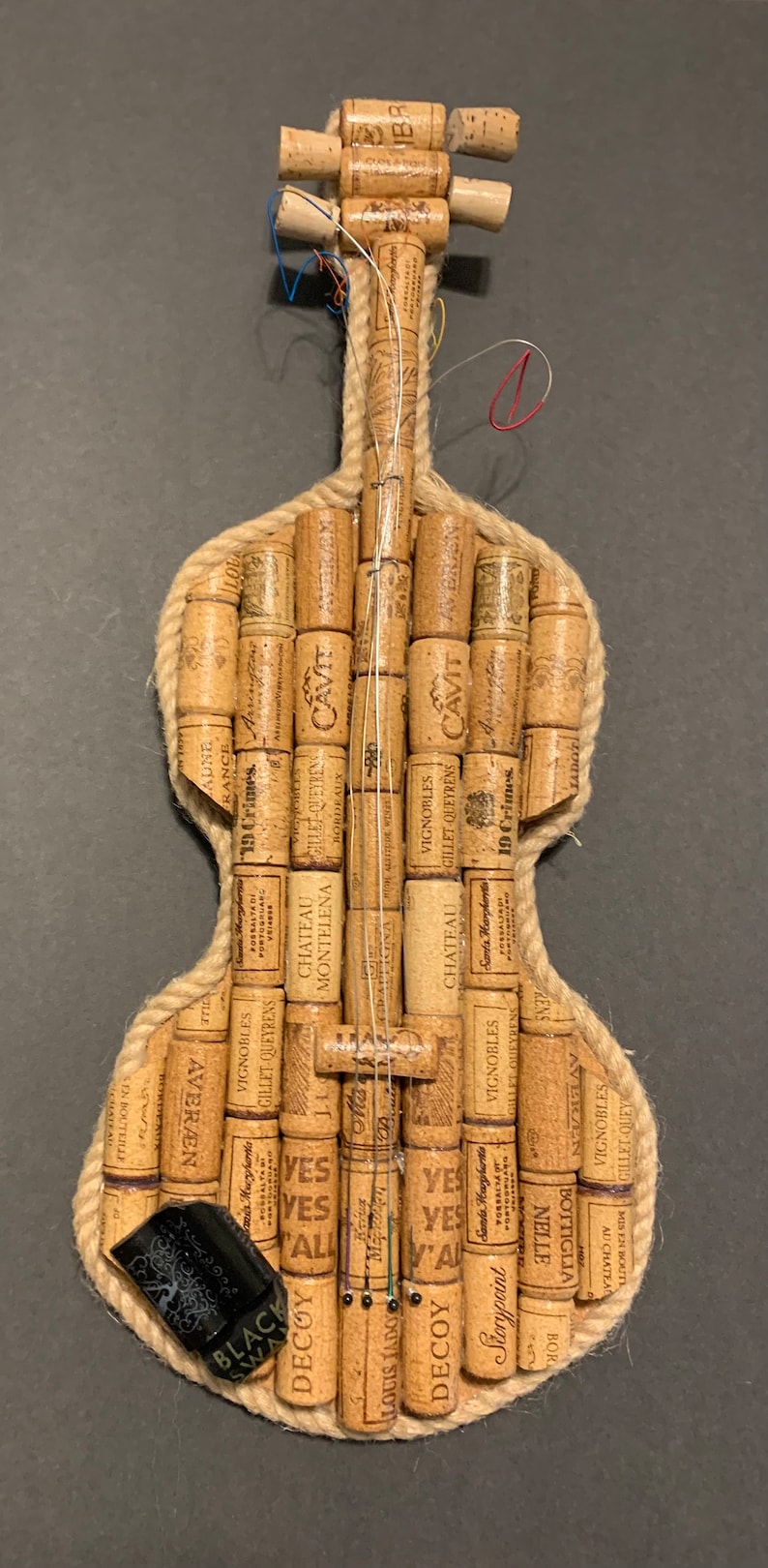 Pu&ograve; includere: Un violino realizzato interamente con tappi di sughero per vino, con un bordo in corda marrone e un'etichetta nera che dice "Black Swan".