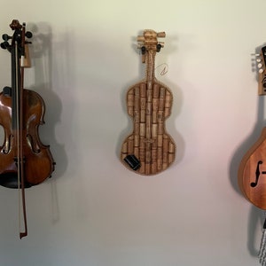 Pu&ograve; includere: Tre strumenti musicali appesi a un muro. Un violino marrone, un violino fatto di tappi di sughero e un mandolino marrone.