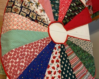 May include: A quilted Christmas tree skirt with a red border. The skirt is made of various fabric panels featuring holiday patterns like candy canes, poinsettias, Christmas lights, and trees. The skirt is designed to wrap around a tree trunk.