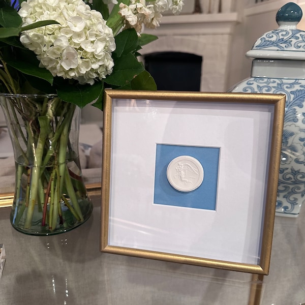 Puede incluir: Un marco dorado con fondo blanco y un paspartú azul. Dentro del paspartú hay una placa de cerámica blanca con un diseño en relieve de un pájaro.