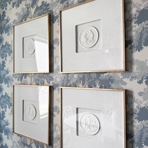 May include: Four framed white relief art pieces with gold trim are displayed on a blue and white patterned wallpaper. Each frame has a square opening with a circular relief sculpture depicting figures.