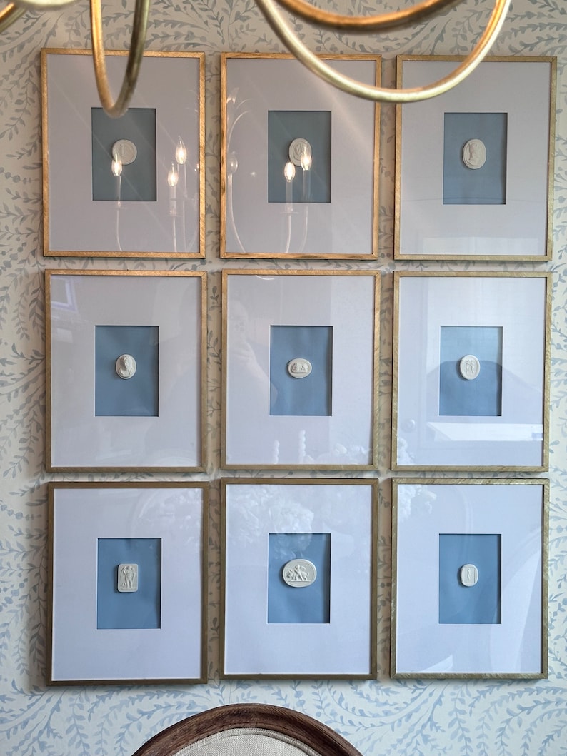 May include: Nine framed antique white relief sculptures with gold frames and light blue mats are displayed on a patterned wallpaper. The sculptures feature classical designs and are arranged in a three-by-three grid.