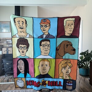 K&ouml;nnte beinhalten: Eine geh&auml;kelte Decke mit den Figuren aus der Zeichentrickserie King of the Hill. Die Decke besteht aus neun Quadraten, die jeweils eine andere Figur zeigen. Die Figuren sind Peggy Hill, Bobby Hill, Dale Gribble, Boomhauer, Hank Hill, Bill Dauterive, Kahn Souphanousinphone und Cotton Hill. Die Decke ist hellblau mit einem wei&szlig;en Rand. Die Worte "King of the Hill" sind in wei&szlig;en Buchstaben auf der Unterseite der Decke geschrieben.