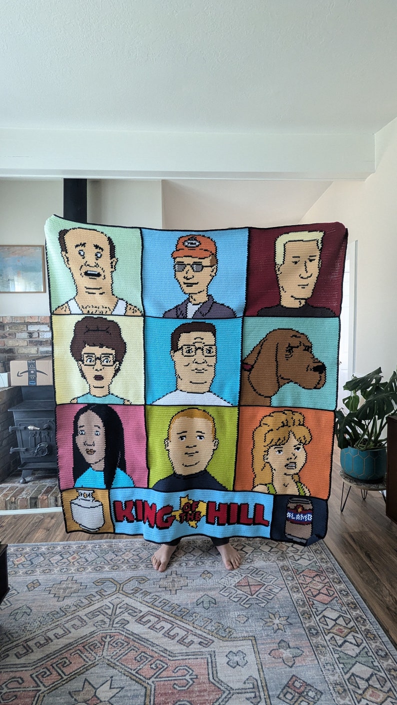 K&ouml;nnte beinhalten: Eine geh&auml;kelte Decke mit den Figuren aus der Zeichentrickserie King of the Hill. Die Decke besteht aus neun Quadraten, die jeweils eine andere Figur zeigen. Die Figuren sind Peggy Hill, Bobby Hill, Dale Gribble, Boomhauer, Hank Hill, Bill Dauterive, Kahn Souphanousinphone und Cotton Hill. Die Decke ist hellblau mit einem wei&szlig;en Rand. Die Worte "King of the Hill" sind in wei&szlig;en Buchstaben auf der Unterseite der Decke geschrieben.