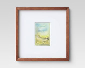 May include: Watercolor landscape art print in a wooden frame. The artwork features a soft, abstract depiction of a landscape with blue, yellow, and green hues. The frame is a warm brown color, and the artwork is matted with a white border.