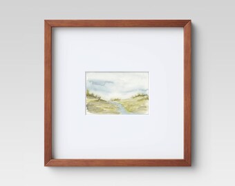 May include: Watercolor landscape art print in a brown wooden frame. The artwork depicts a serene scene with a river flowing through a field, under a cloudy sky. The colors are muted, with shades of green, yellow, and blue.