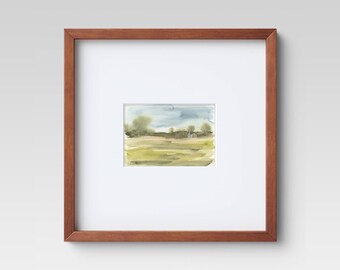May include: A framed watercolor landscape painting. The artwork features a field of green and yellow hues under a blue and gray sky. The painting is set within a white mat and a brown wooden frame.