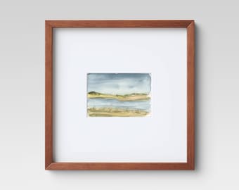 May include: A framed watercolor painting of a landscape with a body of water, hills, and a cloudy sky.
