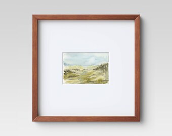 May include: A framed watercolor painting of a landscape with rolling hills, green trees, and a blue sky with white clouds.