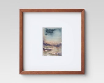 May include: Watercolor landscape painting in a wooden frame. The artwork features a soft, atmospheric scene with muted blues, purples, and beige tones. The painting is framed with a light brown wooden frame and a white mat.