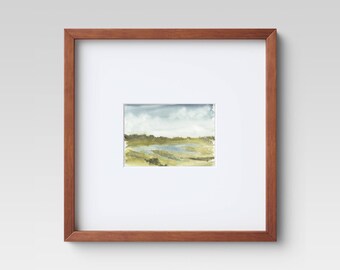 May include: Framed watercolor landscape art. The artwork features a serene scene with a cloudy sky, a body of water, and grassy areas. The painting is set within a white mat and a brown wooden frame.