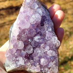 May include: A raw amethyst crystal cluster featuring numerous pointed, translucent purple quartz points. The mineral specimen displays a triangular shape with varying shades of light to deep violet, showcasing natural crystalline formations.
